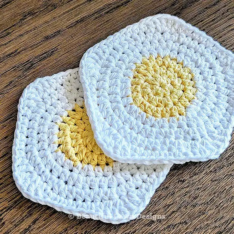Two sunny-side-up egg shaped crochet coasters on a surface