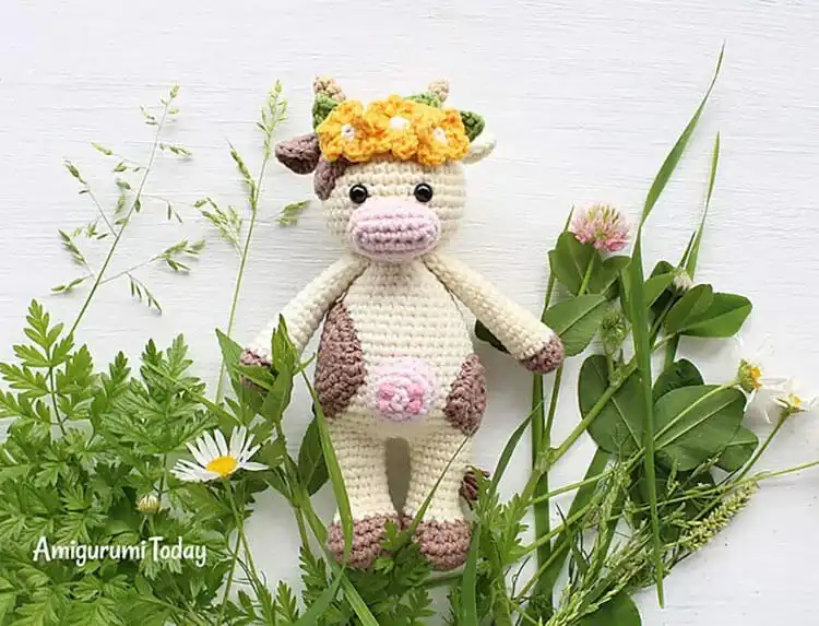 A soft, white crochet Cuddle Me Cow plushie with pink accents, wearing a delicate flower crown on its head.