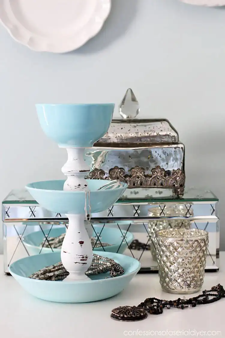A tiered jewelry organizer made from stacked dollar store plates separated by candlesticks, displaying rings and bracelets.