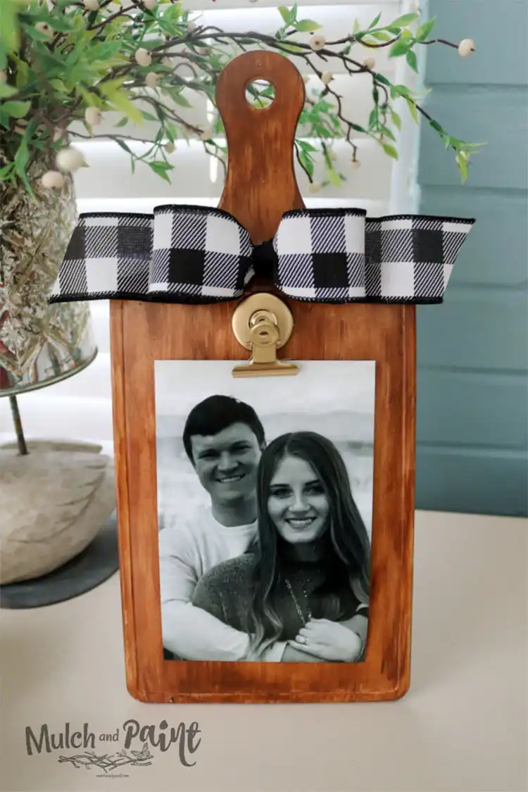 A faux wood cutting board with a clip attached, used as a picture hanger displaying a small photo, hanging on a wall.