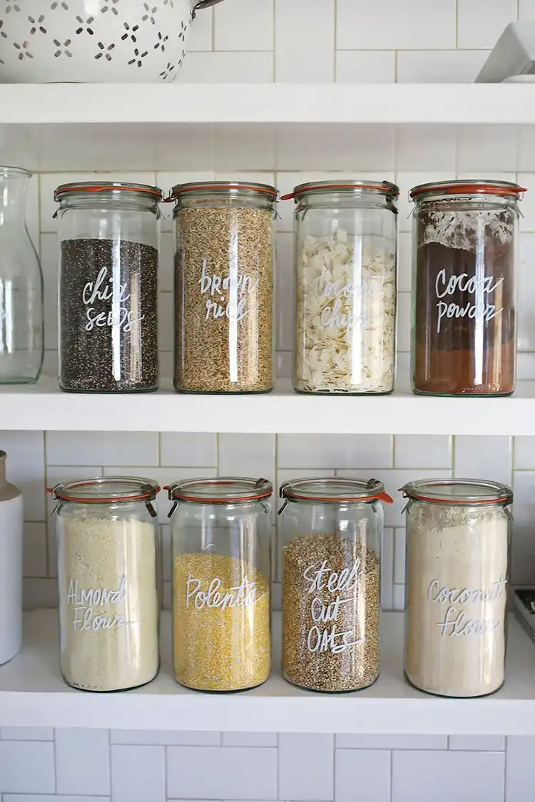 Clear glass jars with neatly hand-lettered labels using white paint pens, organized in a kitchen pantry.