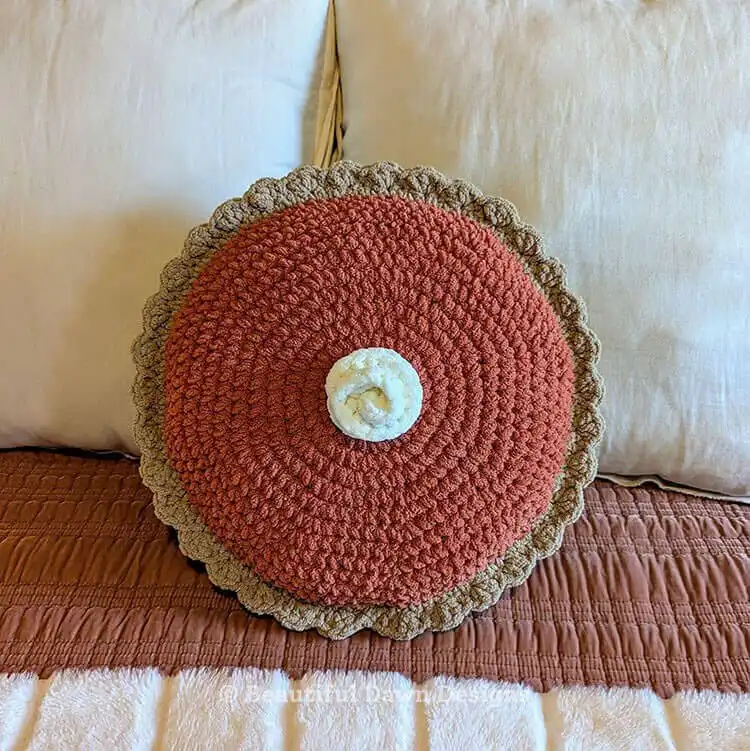 A large, plush crochet pillow designed to look like a slice of pumpkin pie, sitting on a sofa.