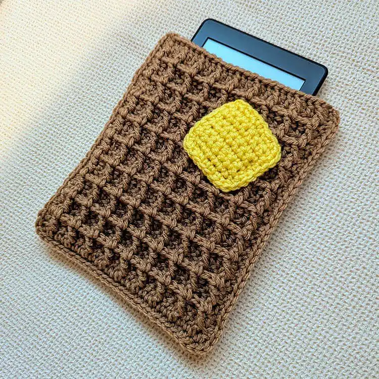 A crocheted e-reader case designed to resemble a buttered waffle, holding an electronic device.