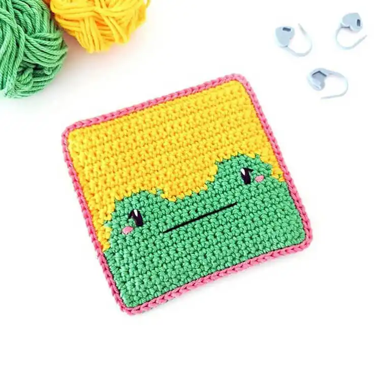 Close-up of a vibrant green crochet frog coaster with white eyes on a wooden table