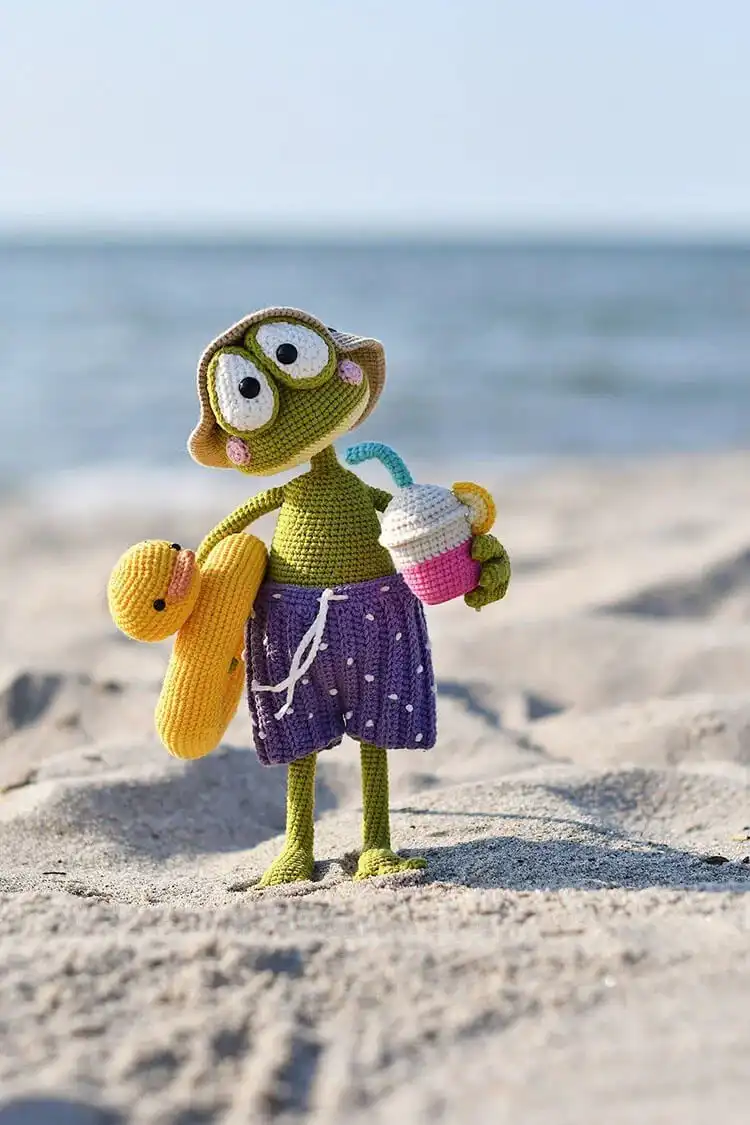 Crochet frog on vacation amigurumi wearing a sun hat, swimwear, and holding a small lemonade, sitting in a rubber ring
