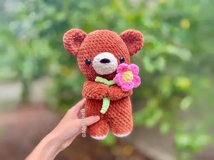 Crochet teddy bear adorned with small colorful crocheted flowers on its head.