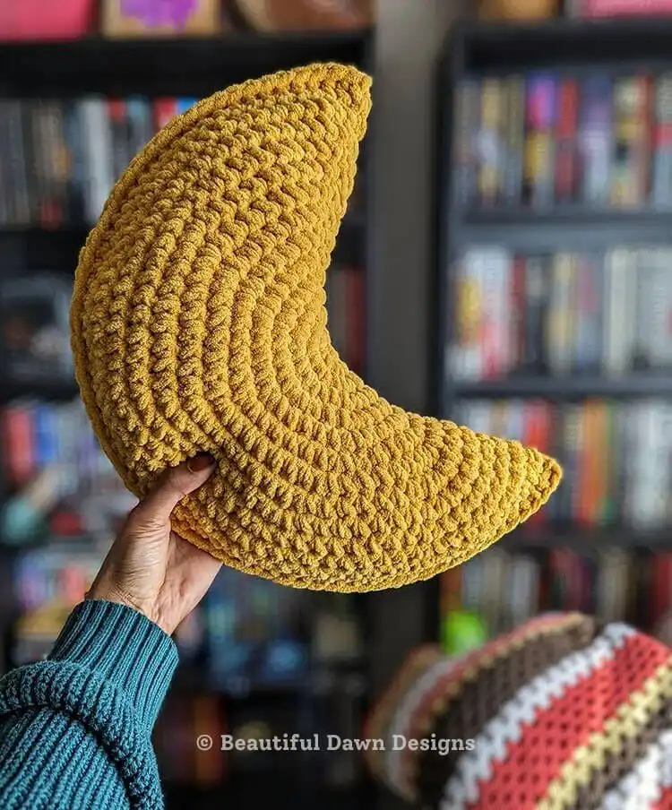 Soft crescent moon crochet pillow in a light cream yarn, resting on a bed