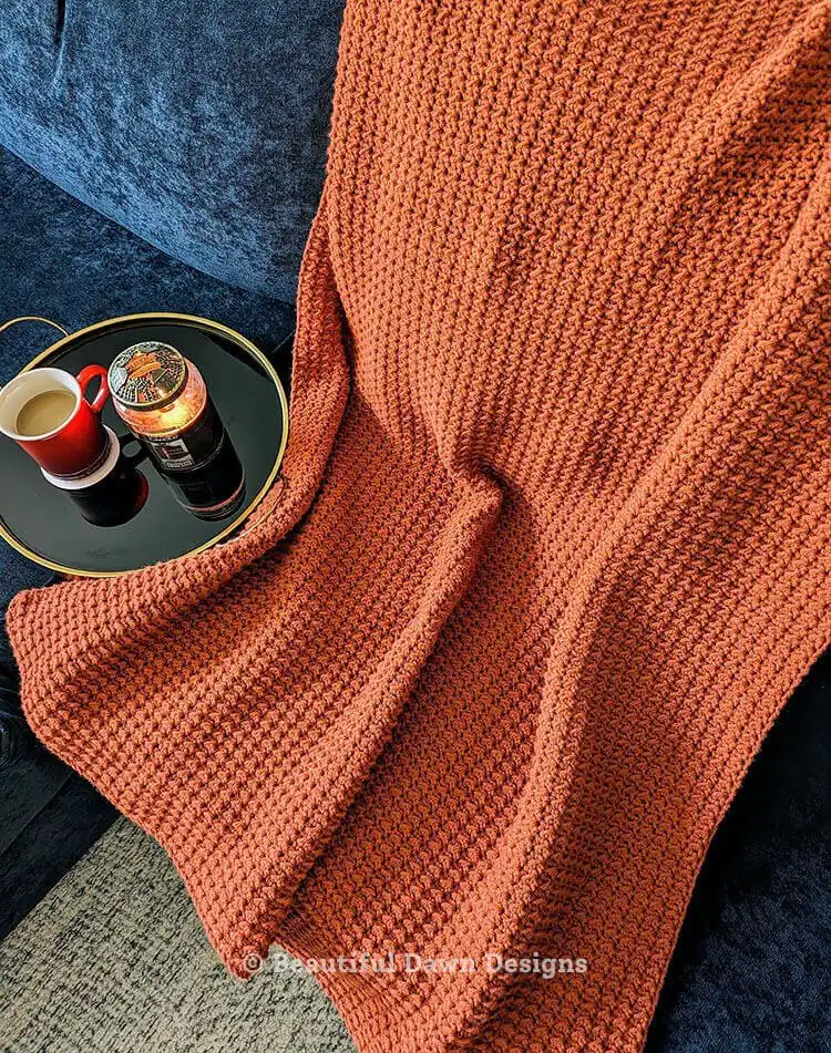 Soft Easy One Row Repeat Crochet Blanket Free Pattern in pumpkin spice yarn draped on a chair, showcasing budget-friendly yarn quality