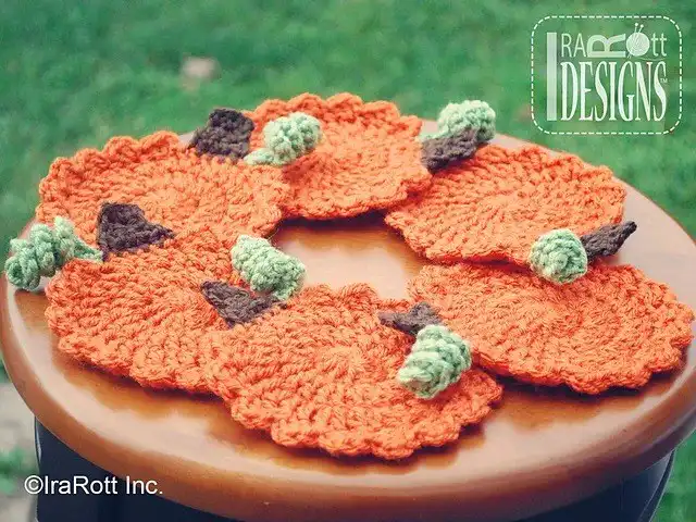 Five crocheted orange Tea Time Pumpkin Coasters with green and brown stems are arranged on a round wooden surface, ready for fall entertaining.