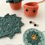Three crocheted coasters shaped like pumpkins in orange, white, and yellow displayed on a wooden surface, with a glass teacup on one.