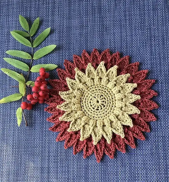 Crocheted coasters with small, colorful pumpkins on the edge, displayed with a glass and crochet tools on a wooden surface, showcasing a 3D Pumpkin Coaster.