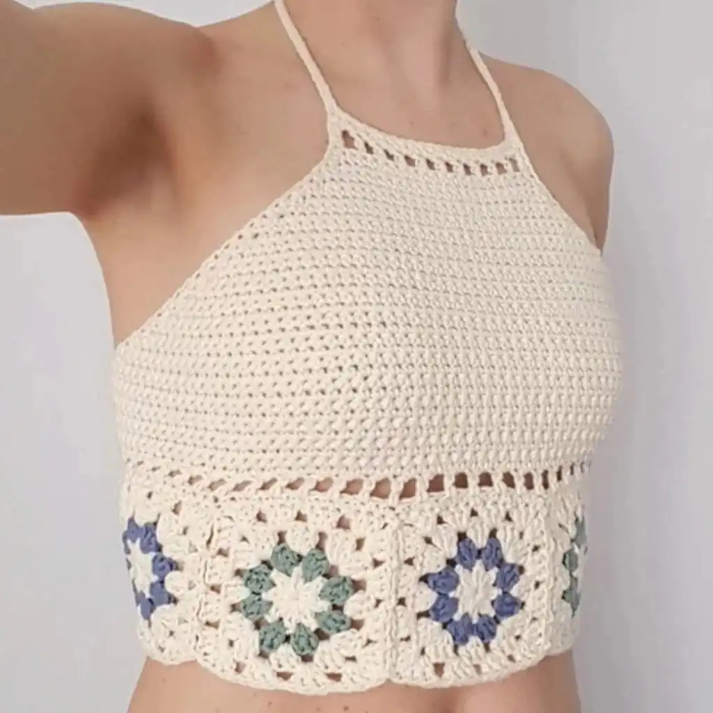 White crochet crop top with intricate patterns and fringe, hanging against green foliage.