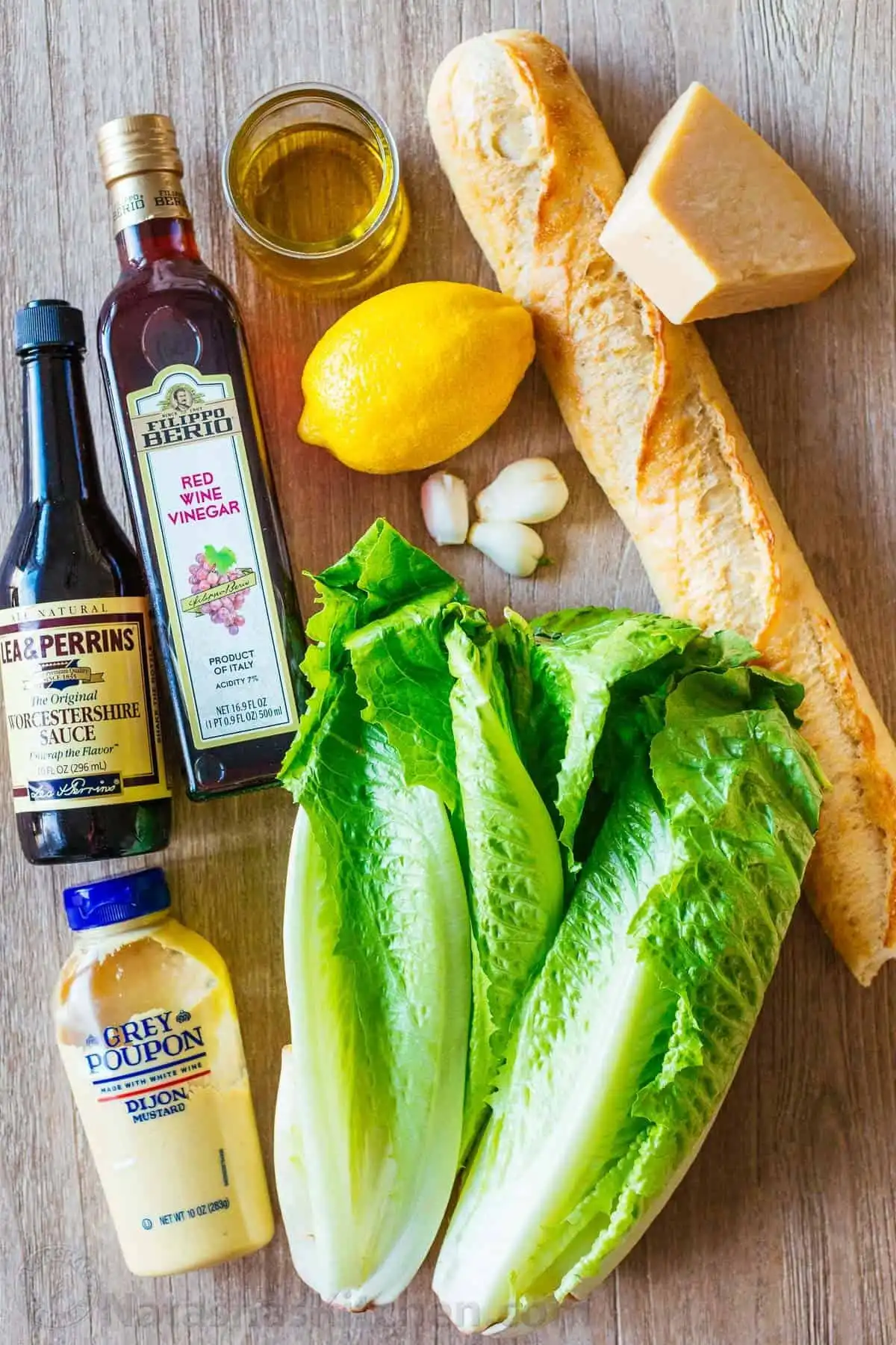Ingredients for classic Caesar Salad including fresh romaine lettuce, a block of Parmesan, baguette for croutons, and dressing components.