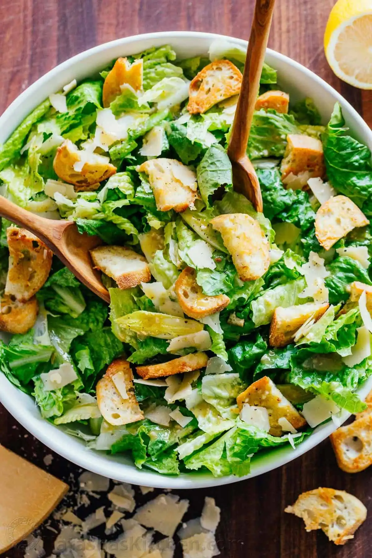 A large bowl of Caesar Salad being tossed with two spoons, showing romaine lettuce, croutons, and Parmesan coated in dressing.