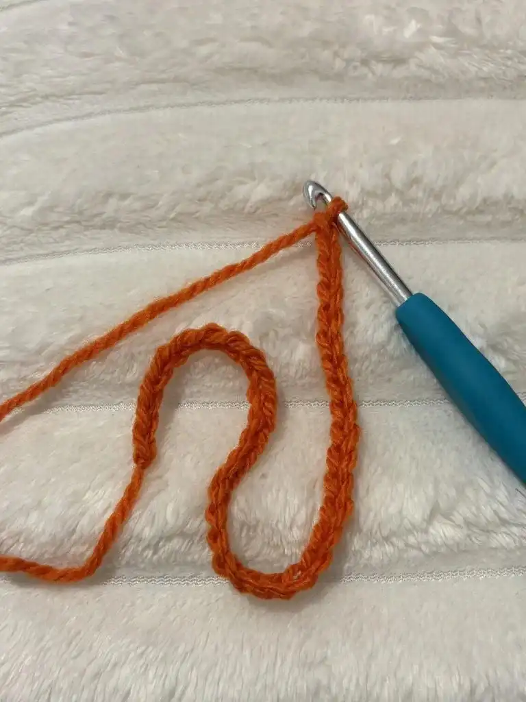 A crochet hook inserted into an orange crocheted chain, demonstrating the initial half double crochet stitches for the pumpkin panel.
