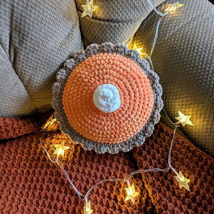Crochet pumpkin pie plushie resting on a rustic wooden chair, showing its full size and inviting texture, as made with the free pattern.