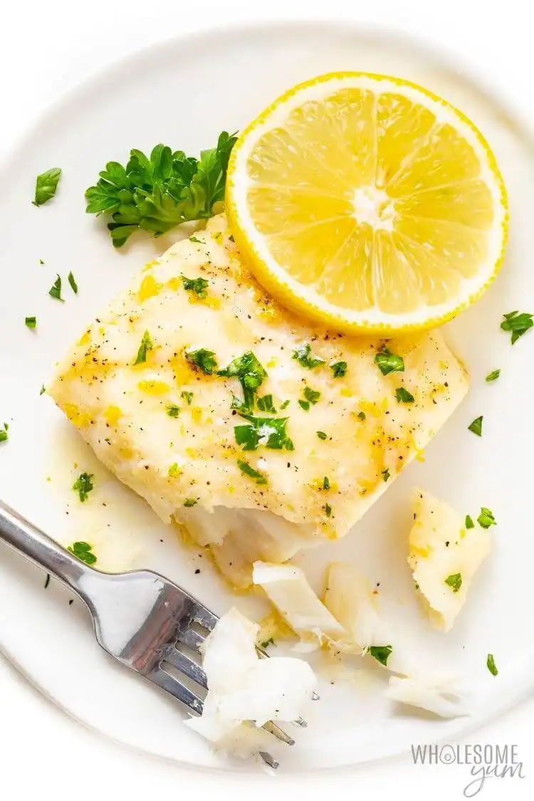 Lemon Baked Cod fillet, flaky and moist, garnished with fresh dill and lemon slices on a white plate.