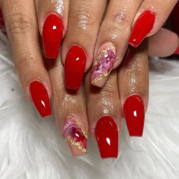 Red acrylic nails with one accent nail featuring a unique marble pattern, making it stand out creatively.
