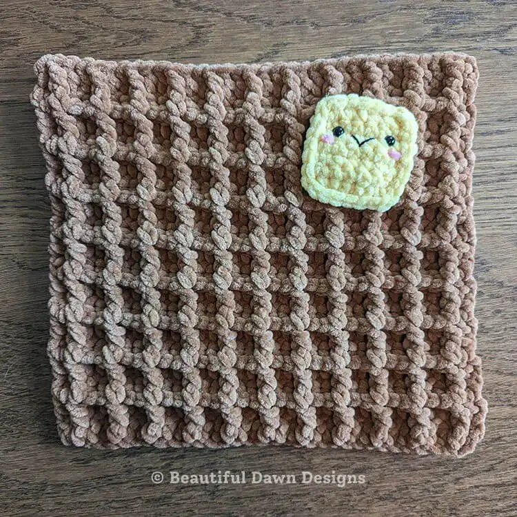 Crocheted yellow butter applique with safety eyes and embroidered details sewn onto one brown waffle panel, part of the Waffle Pillow Free Crochet Pattern.