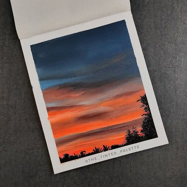 Painting of a dramatic blue and orange sky above trees, capturing vibrant sunrise or sunset colors.