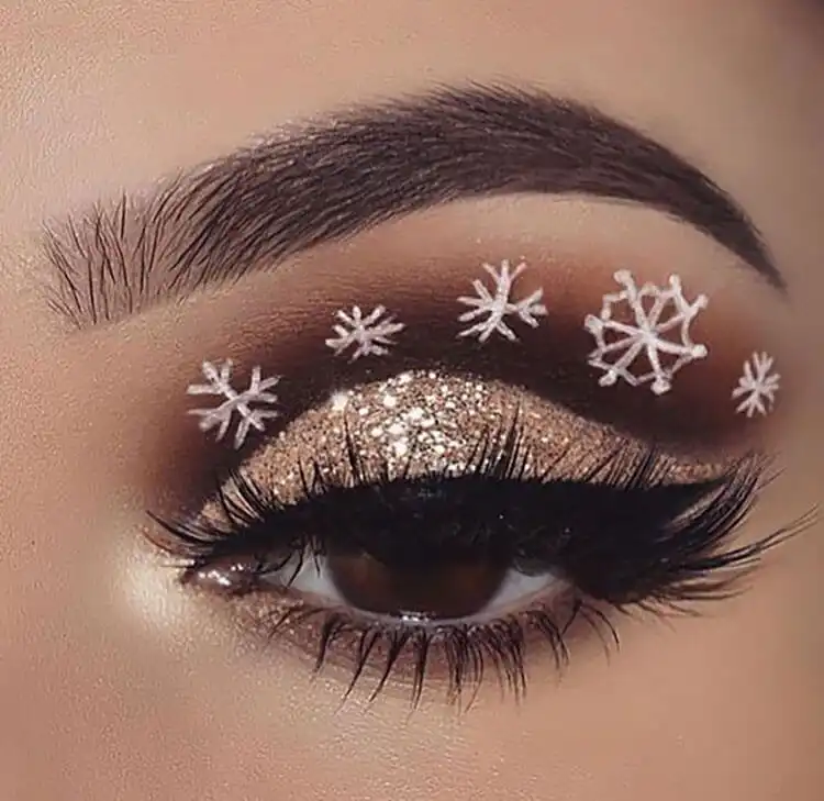 Eye makeup with warm brown eyeshadow, gold glitter, and a delicate white snowflake design at the outer corner.