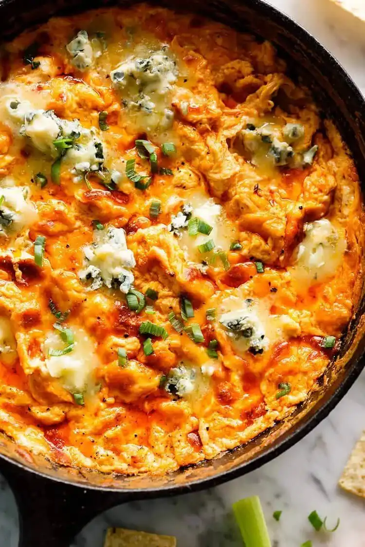 A hot, bubbly buffalo chicken dip in a serving dish, surrounded by tortilla chips and celery sticks.