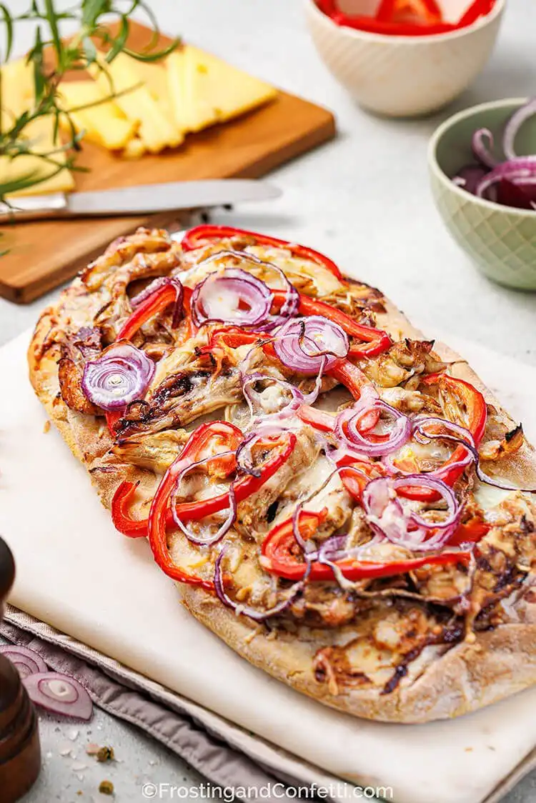 A rustic BBQ chicken flatbread generously topped with shredded rotisserie chicken, barbecue sauce, melted cheese, and fresh red onion slices.